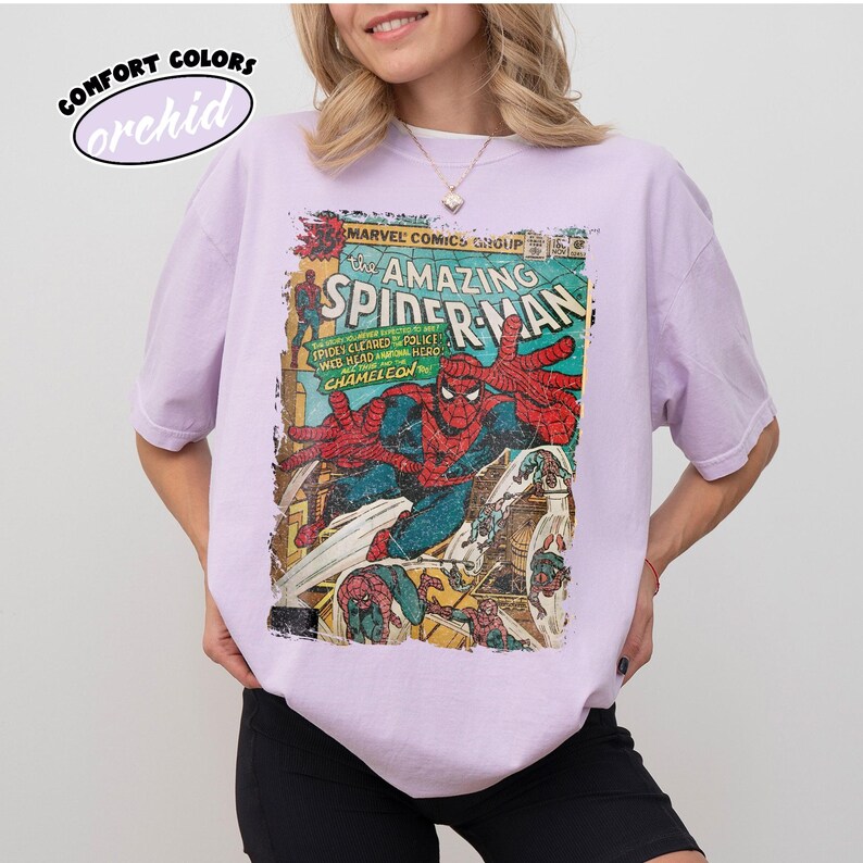May include: A light purple t-shirt featuring a vintage comic book cover of The Amazing Spider-Man. The comic book art shows Spider-Man in action with red and blue colors. The shirt has the words "Comfort Colors" and "orchid" printed on it.