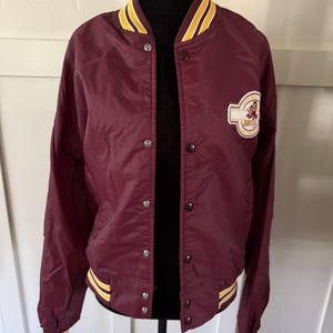 Puede incluir: Chaqueta bomber de color granate con ribete a rayas amarillas y blancas en el cuello, los puños y la cintura. La chaqueta tiene cierre de botones y un parche en el pecho. Una chaqueta de estilo universitario clásico.