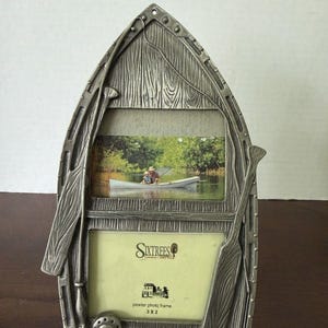 May include: A silver-toned pewter photo frame shaped like a boat, featuring two rectangular openings. The frame is decorated with molded oars and a fishing rod. The bottom opening contains a label reading Sixtrees London New York 3 x 2.