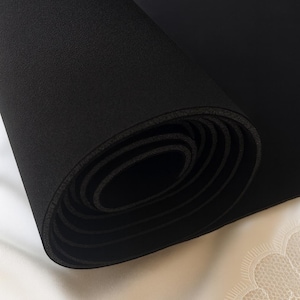 Könnte beinhalten: Eine aufgerollte schwarze Yogamatte. Die Matte besteht aus einem strukturierten Material und ist eng gerollt, wodurch die inneren Schichten sichtbar werden. Der Hintergrund ist eine weiche, weiße Oberfläche mit einem zarten Spitzendesign.