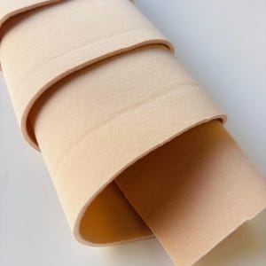 Può includere: Un rotolo di tessuto beige, ben avvolto, con strati visibili. Il tessuto ha una consistenza liscia e un colore neutro, adatto a vari progetti di artigianato o cucito. I bordi del tessuto sono leggermente arrotondati.