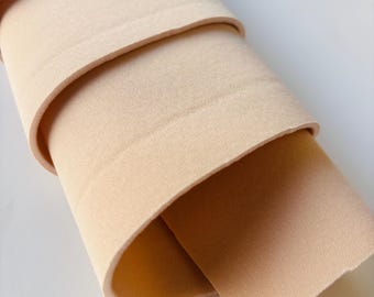 Puede incluir: Un rollo de tela beige, enrollado firmemente, con capas visibles. La tela tiene una textura suave y un color neutro, adecuado para diversos proyectos de manualidades o costura. Los bordes de la tela son ligeramente redondeados.