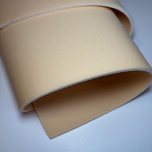 Könnte beinhalten: Nahaufnahme eines gerollten Abschnitts beigen Stoffes. Der Stoff hat eine glatte, matte Oberfläche mit leicht strukturierter Optik. Die Kanten sind sauber und das Material scheint ein weiches, flexibles Textil zu sein. Die Farbe ist ein warmer, neutraler Ton.