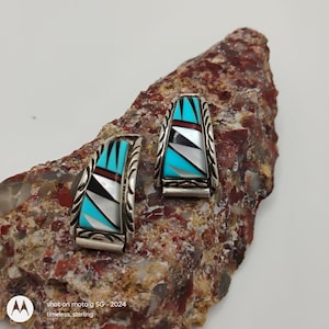 May include: A pair of silver earrings with inlaid turquoise, black, white, and red geometric designs. The earrings are rectangular with curved edges and are displayed on a textured, reddish-brown stone. The text "timeless_sterling" is visible.