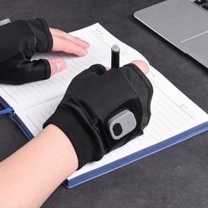 Può includere: Guanti neri senza dita con un pannello di controllo grigio. I guanti vengono utilizzati per scrivere su un taccuino con una penna. Il taccuino è aperto e ha pagine a righe. Un laptop è sullo sfondo.