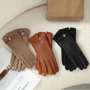Peut inclure: Trois paires de gants en cuir de couleur beige, marron et noir. Chaque paire est ornée d'une lanière avec une boucle dorée. Les gants sont disposés sur une surface blanche, avec un sac à main beige en arrière-plan.