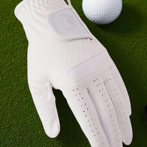 May include: A white golf glove with a logo on the wrist strap and small perforations on the palm. A white golf ball is next to the glove on a green surface. The glove is made of a smooth material.