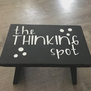 May include: A small, black wooden stool with the words "the THINKING spot" in white script. White dots are scattered around the text. The stool has four legs and is designed for sitting.