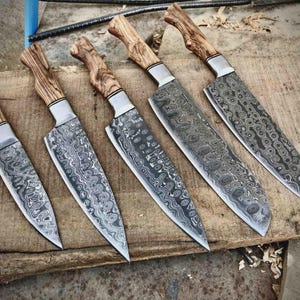May include: Five handcrafted knives with patterned steel blades and wooden handles. The blades have a unique, swirling design. The handles are made of light-colored wood. The knives are arranged on a wooden surface.