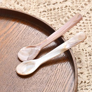 Op de afbeelding: Twee decoratieve lepels met parelmoeren handvatten en kommen liggen op een houten dienblad. Eén lepel is lichtroze en de andere wit. De lepels zijn waarschijnlijk bedoeld om te serveren of tentoon te stellen.