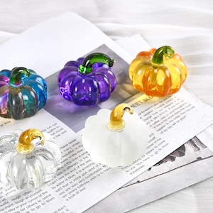 May include: A collection of decorative glass pumpkins in various colors, including blue, purple, orange, and clear. Each pumpkin has a stem and is placed on a newspaper background. A fall-themed decorative item.