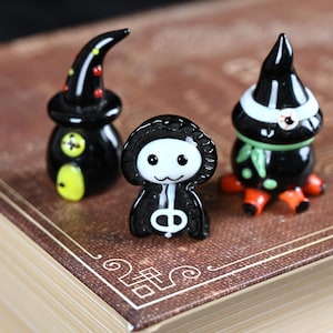 以下が含まれることがあります： ハロウィーンをテーマにした3つのガラス製フィギュア。フィギュアには、魔女の帽子、フード付きのスケルトン、目のある生き物が含まれています。フィギュアは黒色でカラフルなアクセントがあり、茶色の表面に置かれています。