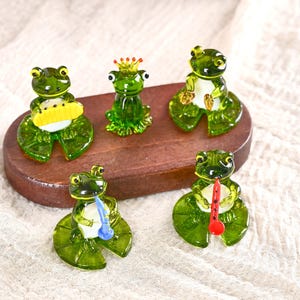 以下が含まれることがあります： 木製の台座に置かれた、5匹の緑色のガラス製カエルの置物。それぞれ異なるポーズをしており、楽器を持っているもの、王冠をかぶっているもの、黄色いものを持っているものなどがあります。カエルは緑色のスイレンの葉の上に置かれています。