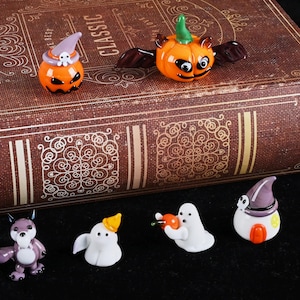 Może przedstawiać: Kolekcja szklanych figurek o tematyce Halloween. Figurki obejmują dynie, duchy i fioletowego wilka. Dynie mają skrzydła i kapelusze czarownic. Figurki znajdują się na brązowej książce z napisem "CLASSIC" na grzbiecie.