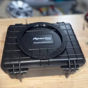 May include: A black Apache 1800 Rugged Mobility case with a textured, circular insert. The case has a rectangular shape with a grid pattern and secure latches. The case is designed for rugged use.