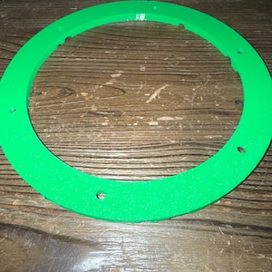 Puede incluir: Un objeto circular verde brillante con una superficie texturizada y varios agujeros espaciados uniformemente. El objeto tiene forma de anillo, posiblemente un componente de un art&iacute;culo m&aacute;s grande. El color es un verde vibrante y s&oacute;lido.