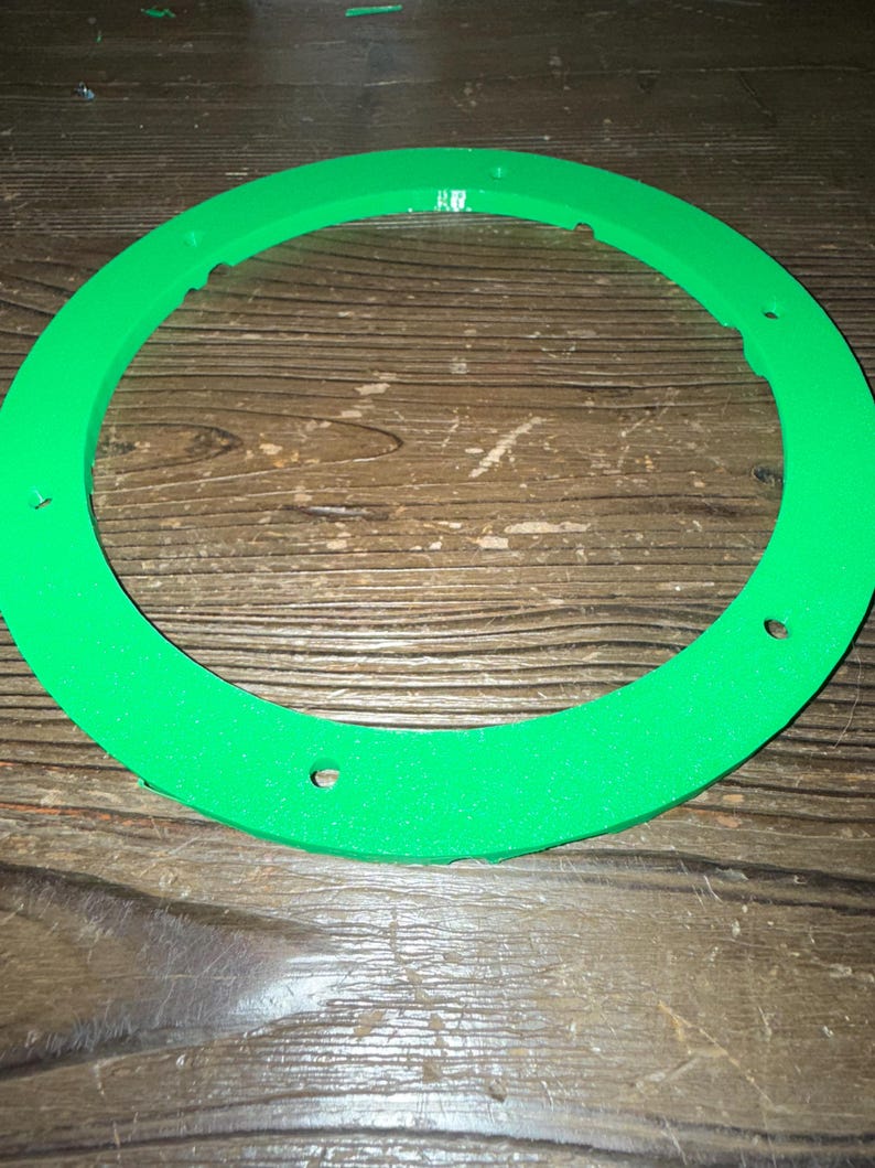 Puede incluir: Un objeto circular verde brillante con una superficie texturizada y varios agujeros espaciados uniformemente. El objeto tiene forma de anillo, posiblemente un componente de un art&iacute;culo m&aacute;s grande. El color es un verde vibrante y s&oacute;lido.