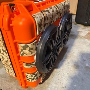 Puede incluir: Un altavoz port&aacute;til naranja con dos altavoces negros y detalles de camuflaje. El altavoz tiene un asa y est&aacute; dise&ntilde;ado para ser resistente y duradero. El patr&oacute;n de camuflaje es marr&oacute;n, beige y verde.