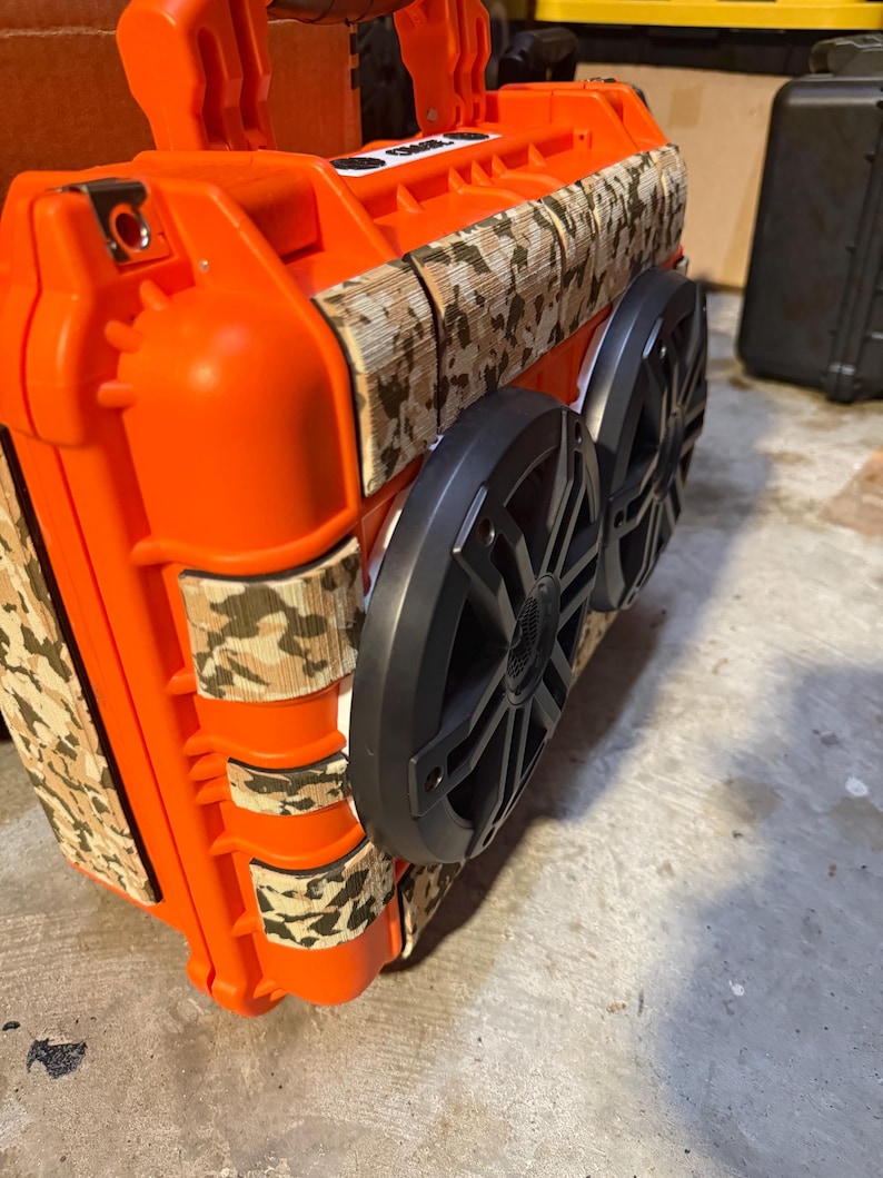 Puede incluir: Un altavoz port&aacute;til naranja con dos altavoces negros y detalles de camuflaje. El altavoz tiene un asa y est&aacute; dise&ntilde;ado para ser resistente y duradero. El patr&oacute;n de camuflaje es marr&oacute;n, beige y verde.