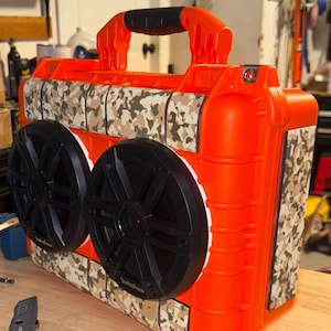 Puede incluir: Un altavoz port&aacute;til naranja con dos grandes altavoces negros y un patr&oacute;n de camuflaje. El altavoz tiene un asa para transportarlo y est&aacute; dise&ntilde;ado para uso en exteriores. Est&aacute; sobre una superficie de madera.