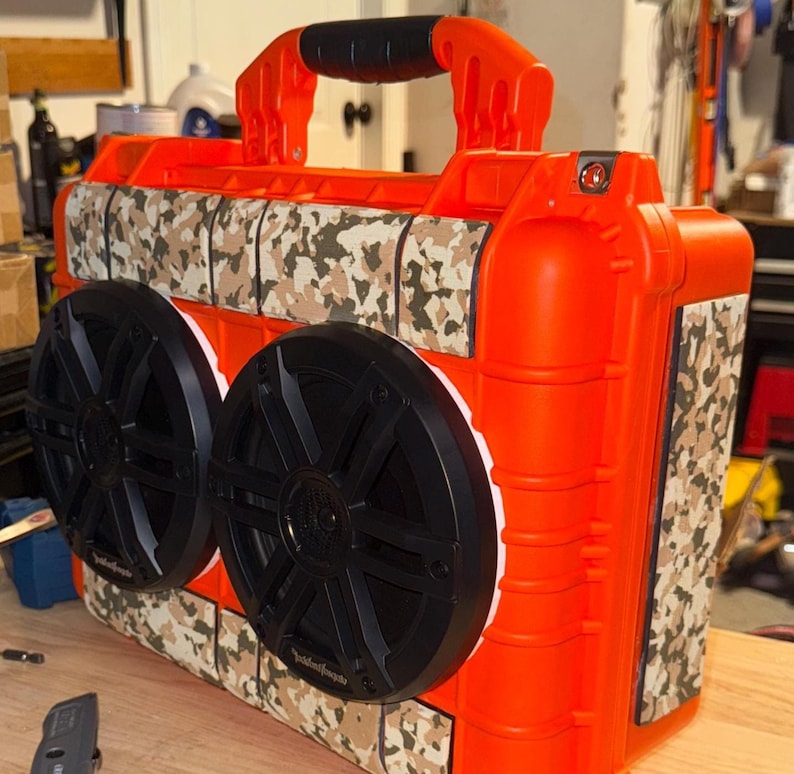 Puede incluir: Un altavoz port&aacute;til naranja con dos grandes altavoces negros y un patr&oacute;n de camuflaje. El altavoz tiene un asa para transportarlo y est&aacute; dise&ntilde;ado para uso en exteriores. Est&aacute; sobre una superficie de madera.