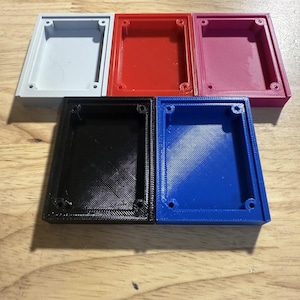 May include: Six rectangular plastic boxes in various colours: grey, red, pink, black, and blue. Each box has a recessed interior and four small holes in the corners. The boxes are arranged on a wooden surface.