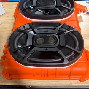 以下が含まれることがあります： オレンジ色の筐体に、メッシュセンターを備えた2つの黒いPolk DBシリーズスピーカーが取り付けられています。スピーカーは黒いフレームで、楕円形をしています。筐体は長方形で、表面はテクスチャ加工されています。