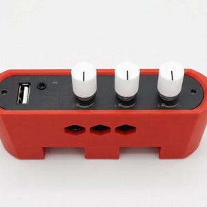 Puede incluir: Un dispositivo electr&oacute;nico rojo con tres perillas blancas y un puerto USB. El dispositivo tiene forma rectangular con bordes redondeados y peque&ntilde;as aberturas. Las perillas son cil&iacute;ndricas con una l&iacute;nea negra en la parte superior.