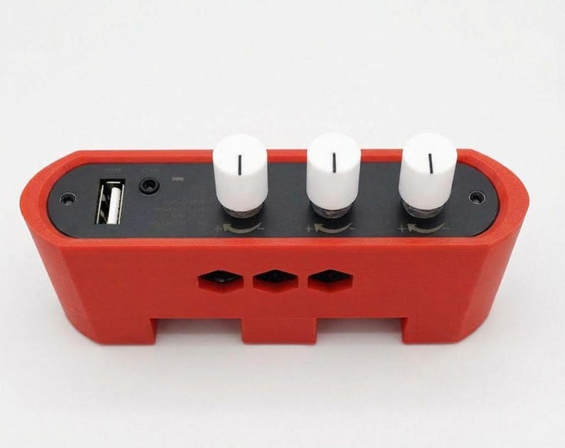 Puede incluir: Un dispositivo electr&oacute;nico rojo con tres perillas blancas y un puerto USB. El dispositivo tiene forma rectangular con bordes redondeados y peque&ntilde;as aberturas. Las perillas son cil&iacute;ndricas con una l&iacute;nea negra en la parte superior.