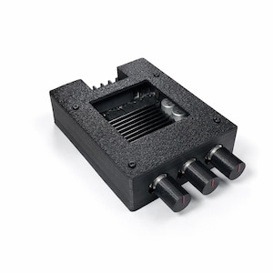 Op de afbeelding: Een zwart elektronisch apparaat met een getextureerd oppervlak en drie knoppen met rode accenten. Het apparaat heeft een rechthoekige vorm met een verzonken bovendeel en een reeks horizontale lijnen. De knoppen zijn op een rij geplaatst.