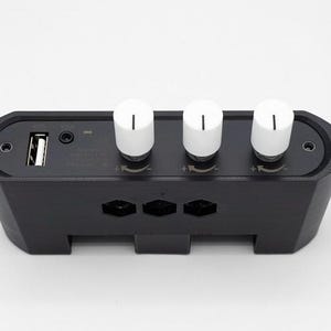 Puede incluir: Un dispositivo electr&oacute;nico negro con tres perillas blancas y un puerto USB. El dispositivo tiene forma rectangular con bordes redondeados y cortes geom&eacute;tricos. Las perillas son cil&iacute;ndricas con una l&iacute;nea negra en la parte superior. Probablemente una mesa de mezclas.