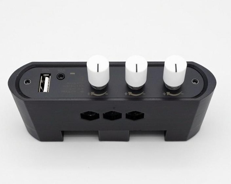 Puede incluir: Un dispositivo electr&oacute;nico negro con tres perillas blancas y un puerto USB. El dispositivo tiene forma rectangular con bordes redondeados y cortes geom&eacute;tricos. Las perillas son cil&iacute;ndricas con una l&iacute;nea negra en la parte superior. Probablemente una mesa de mezclas.