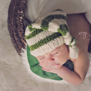 Puede incluir: Un bebé recién nacido con un gorro de punto a rayas verdes y blancas con un nudo en la parte superior. El bebé está durmiendo en una cesta con una manta blanca.