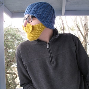 May include: A person wearing a blue knitted hat and a gray sweater with a yellow knitted beard.