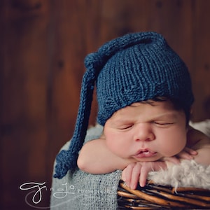 Puede incluir: Un bebé recién nacido con un gorro azul de punto con una punta larga y puntiaguda, durmiendo en una cesta de mimbre forrada con una manta blanca y esponjosa.