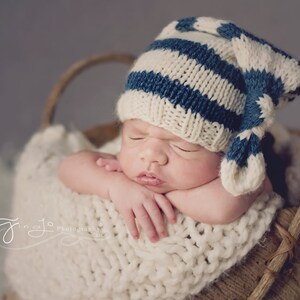 Puede incluir: Un bebé recién nacido con un gorro de punto azul y blanco a rayas con una punta larga y puntiaguda, durmiendo en una cesta de punto blanca.