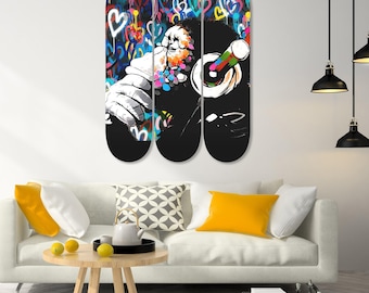 Op de afbeelding: Triptiek wandkunst met een graffiti-achtige afbeelding van een figuur met een koptelefoon. Het kunstwerk wordt weergegeven op drie skateboard decks. De achtergrond is gevuld met kleurrijke harten en abstracte vormen. De hand van de figuur houdt een kleurrijk object vast.