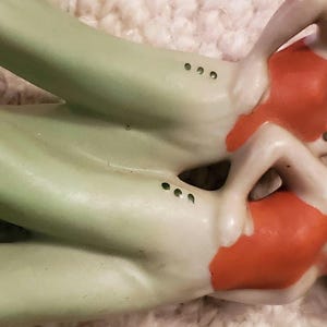 May include: Two porcelain figurines of mermaids. The mermaids have green tails and wear orange tops. The figurines are lying down, with their arms bent. The background is a light, textured surface.