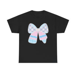 May include: Black t-shirt with a large bow design in pastel colours. The bow is made up of pink, blue, and white patchwork patterns, including gingham and floral prints. The bow is outlined with a white border.