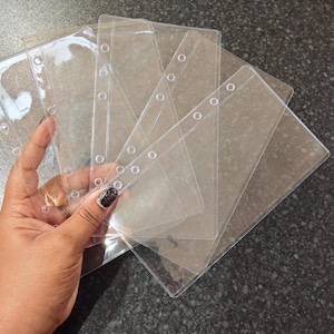 May include: A hand holding a stack of clear, rectangular plastic binder pockets. Each pocket has a series of reinforced holes along one side. The pockets are transparent, allowing visibility of contents. The background is a dark, textured surface.