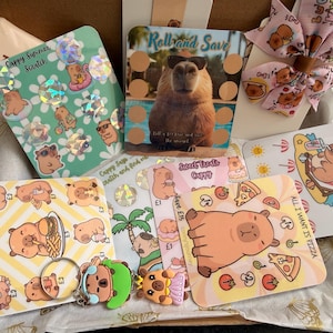 May include: A selection of illustrated cards and accessories with cartoon capybara designs. The cards feature scratch-off games, pizza, and summer themes. A pink bow with capybara graphics is also included in the assortment.