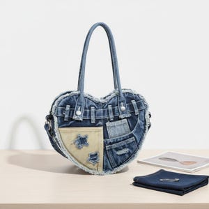 May include: A heart-shaped denim handbag with a patchwork design. The bag features a light blue denim color with various denim patches and a beige accent. It has two handles and a shoulder strap. A denim napkin with a safety pin and a magazine are also visible.