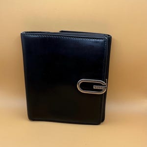 Puede incluir: Una cartera plegable de cuero negro con acabado liso y un cierre de metal plateado en forma de G en la parte frontal. La palabra GUCCI está grabada en letras mayúsculas pequeñas en la barra horizontal inferior del herraje metálico.