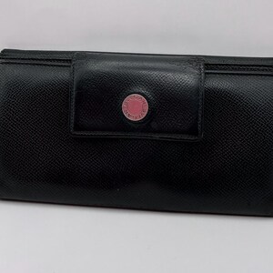 May include: A black leather wallet with a textured surface and a rectangular flap closure. The centre of the flap features a circular pink button accent with the text BVLGARI embossed around the perimeter in a metallic silver tone.