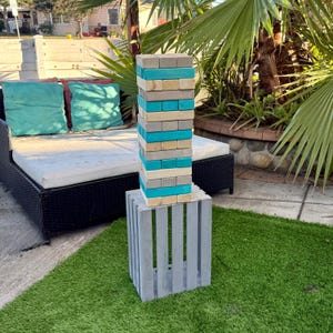 May include: A tall tower game made of wooden blocks in shades of blue, gray, and beige. The tower is stacked on a gray wooden crate, set on a green lawn. The game is outdoors near a patio set with blue and red pillows.