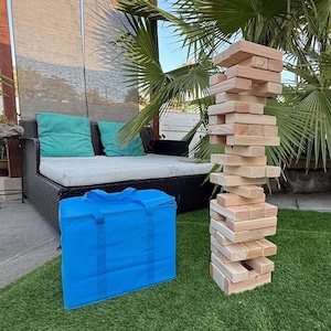 May include: A large outdoor wooden block stacking game, with a blue carrying case. The game is set up on a green lawn, with a black outdoor sofa and palm trees in the background. The blocks are made of natural wood.
