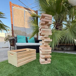 May include: A wooden Jenga game tower with a wooden crate on a green lawn. The Jenga tower is made of rectangular wooden blocks stacked in alternating directions. The crate is rectangular with a handle on each side.