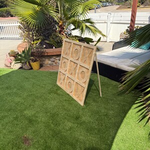 May include: A wooden tic-tac-toe game board with the letters "X" and "O" carved into the surface. The game is propped up on a wooden stand on a patch of green artificial turf. The background includes a palm tree and a white fence.
