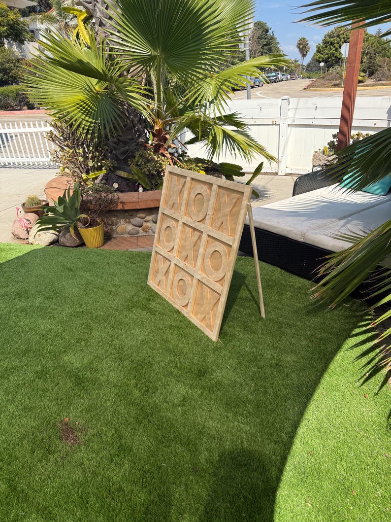 May include: A wooden tic-tac-toe game board with the letters "X" and "O" carved into the surface. The game is propped up on a wooden stand on a patch of green artificial turf. The background includes a palm tree and a white fence.