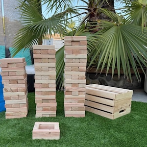 May include: A wooden block stacking game with multiple towers of rectangular blocks, a small square block, and a wooden crate. The blocks are light brown and arranged on a green surface, suggesting outdoor play.